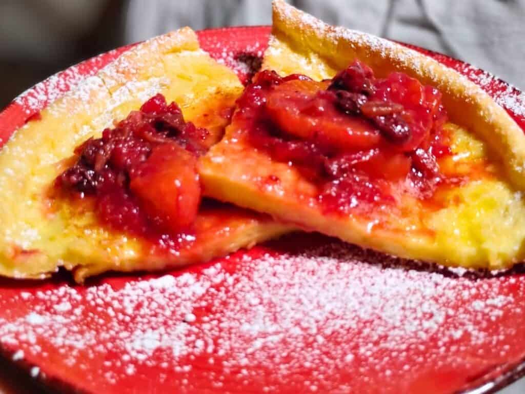 photo shows dutch baby pancake slices topped with a cranberry orange sauce on a red plate, sprinkled with powder sugar.