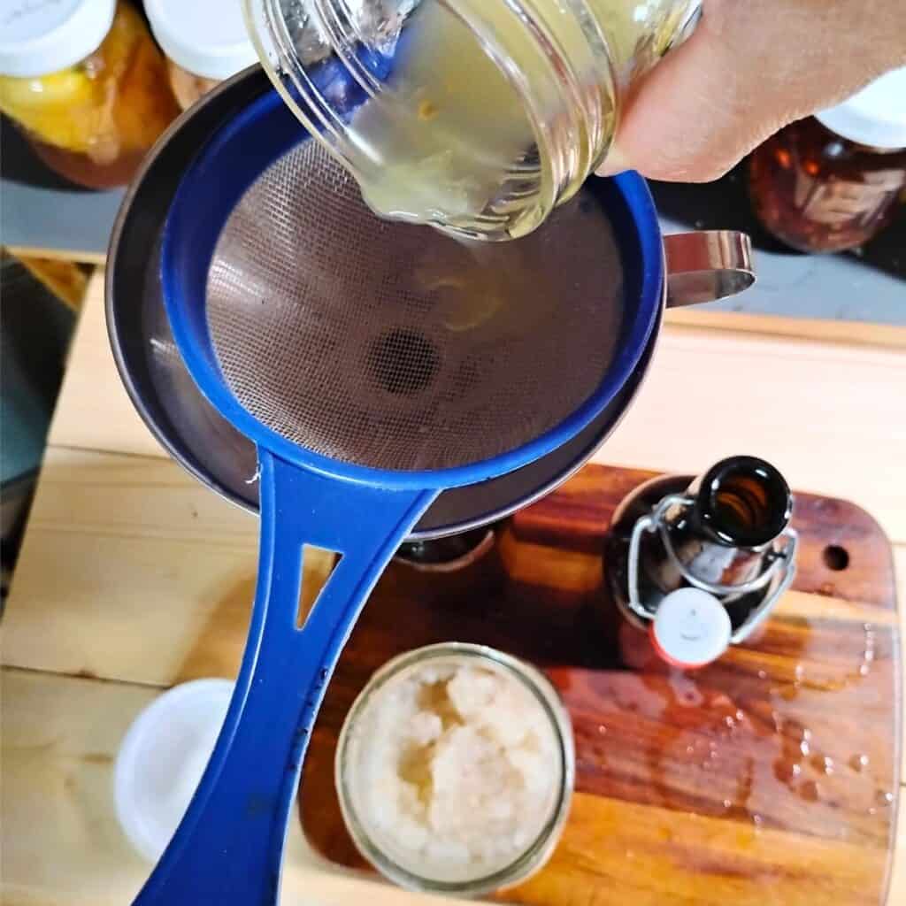 a hand is holding a jar with lemon juice and pouring it thorough the strainer and funnel into the bottle.
