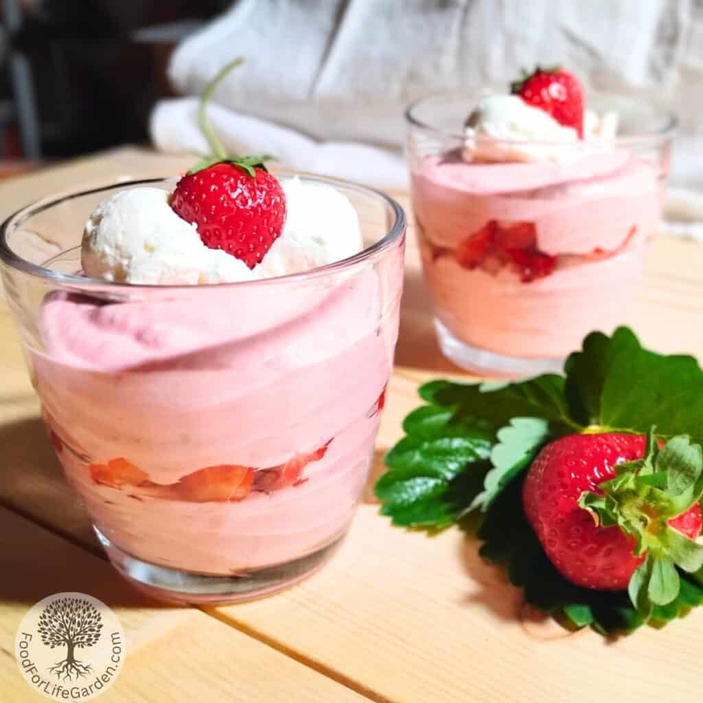photo of two jars full of German style quark cheese mousse with a layer of strawberries and decorated with whipped cream and strawberries. There is a strawberry leaf and a strawberry next to it.