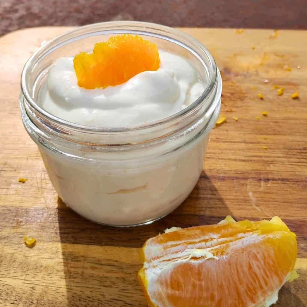 glass bowl full of quark cheese dessert with an orange slice for decoration and a piece of orange in front of it.