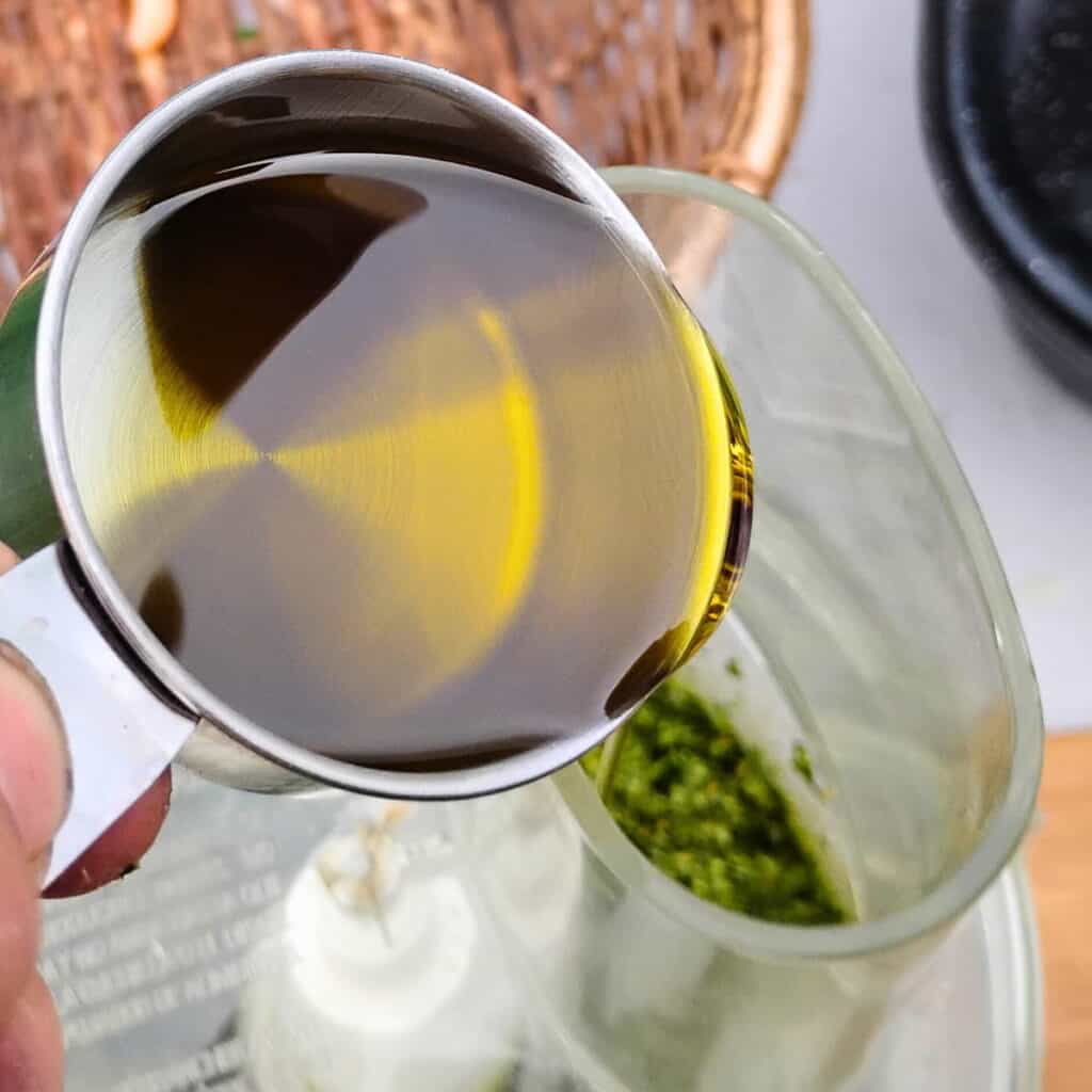 olive oil is getting poured into the running processor with foraged wild greens pesto ingredients.