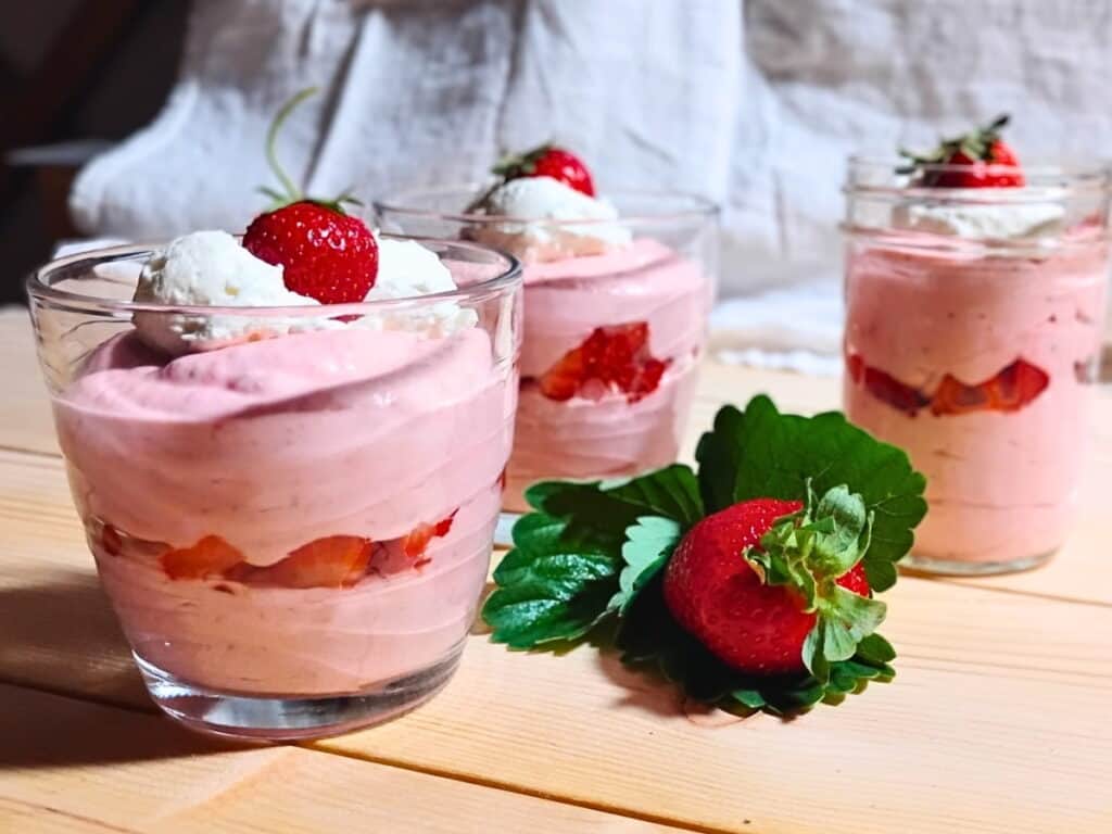 three serving glasses full of strawberry quark cheese mousse, layered with strawberries and topped with strawberries and whipped cream.