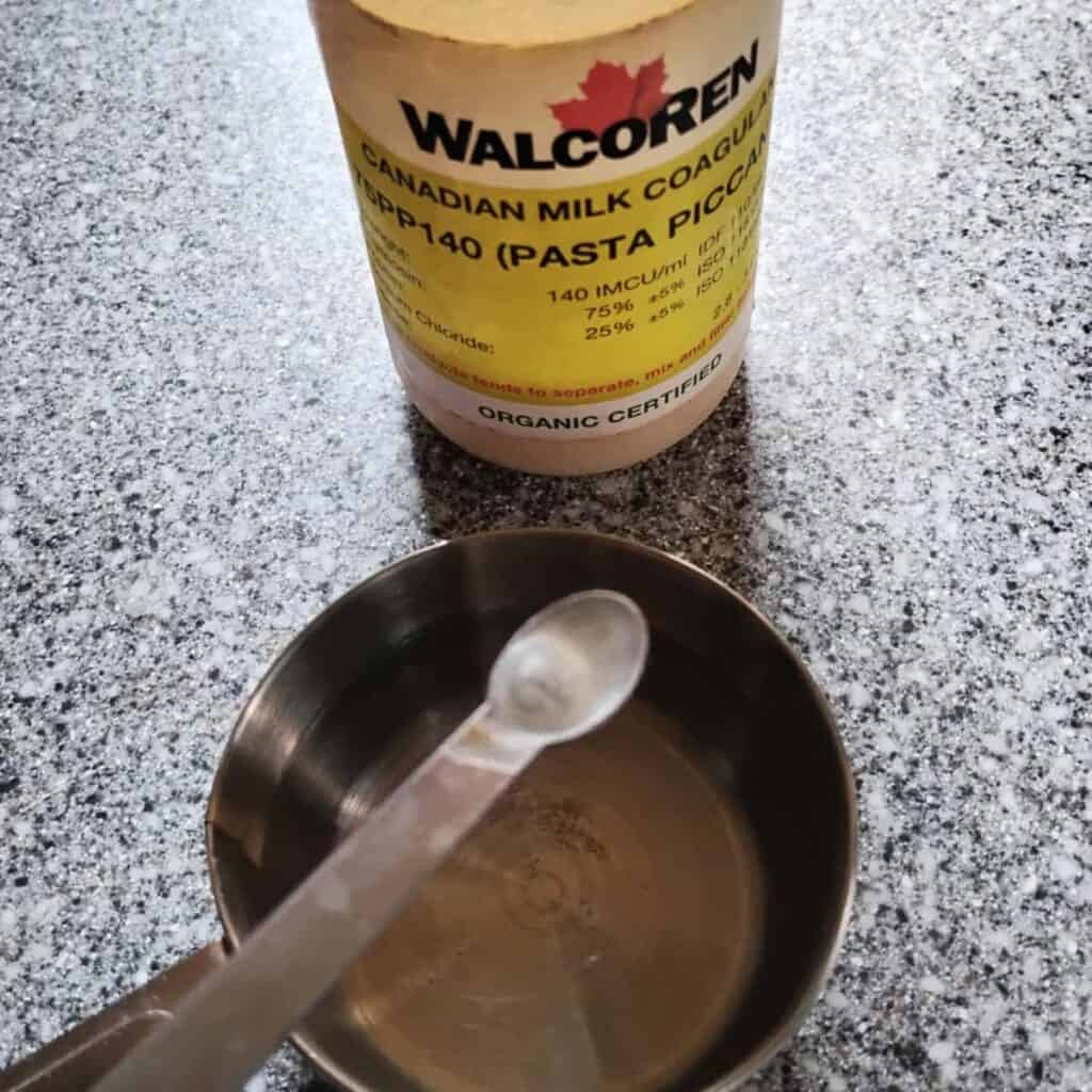 bottle of kid rennet and a spoon measuring out a dose into a measuring cup with water.