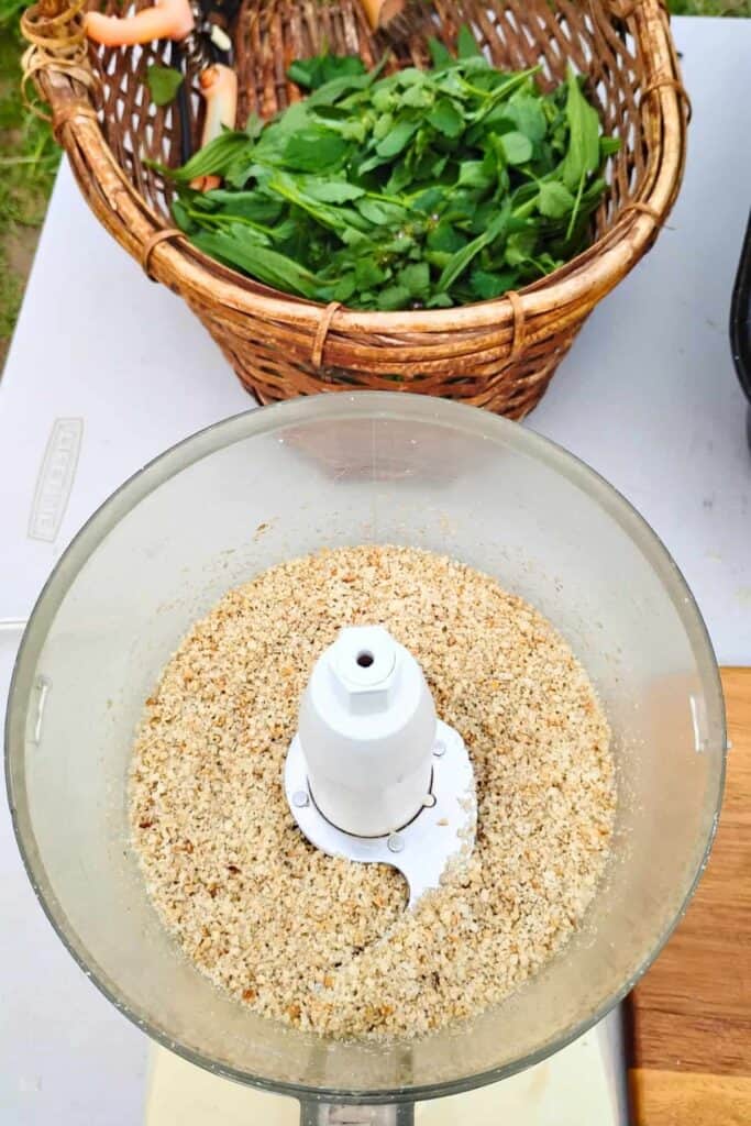 processed sunflower seeds, garlic and cheese processed to resemble fine crumbs. there are foraged wild greens in a basket behind it.