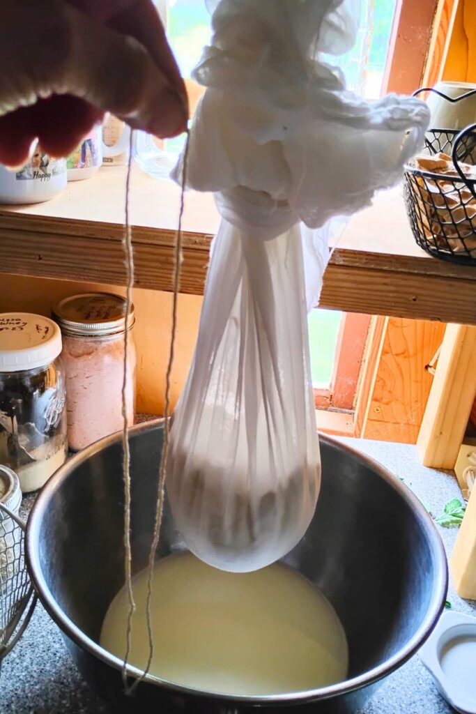 A bag of cheese hanging from a hook and dripping whey into a bowl. a hand is holding a piece of string next to it.