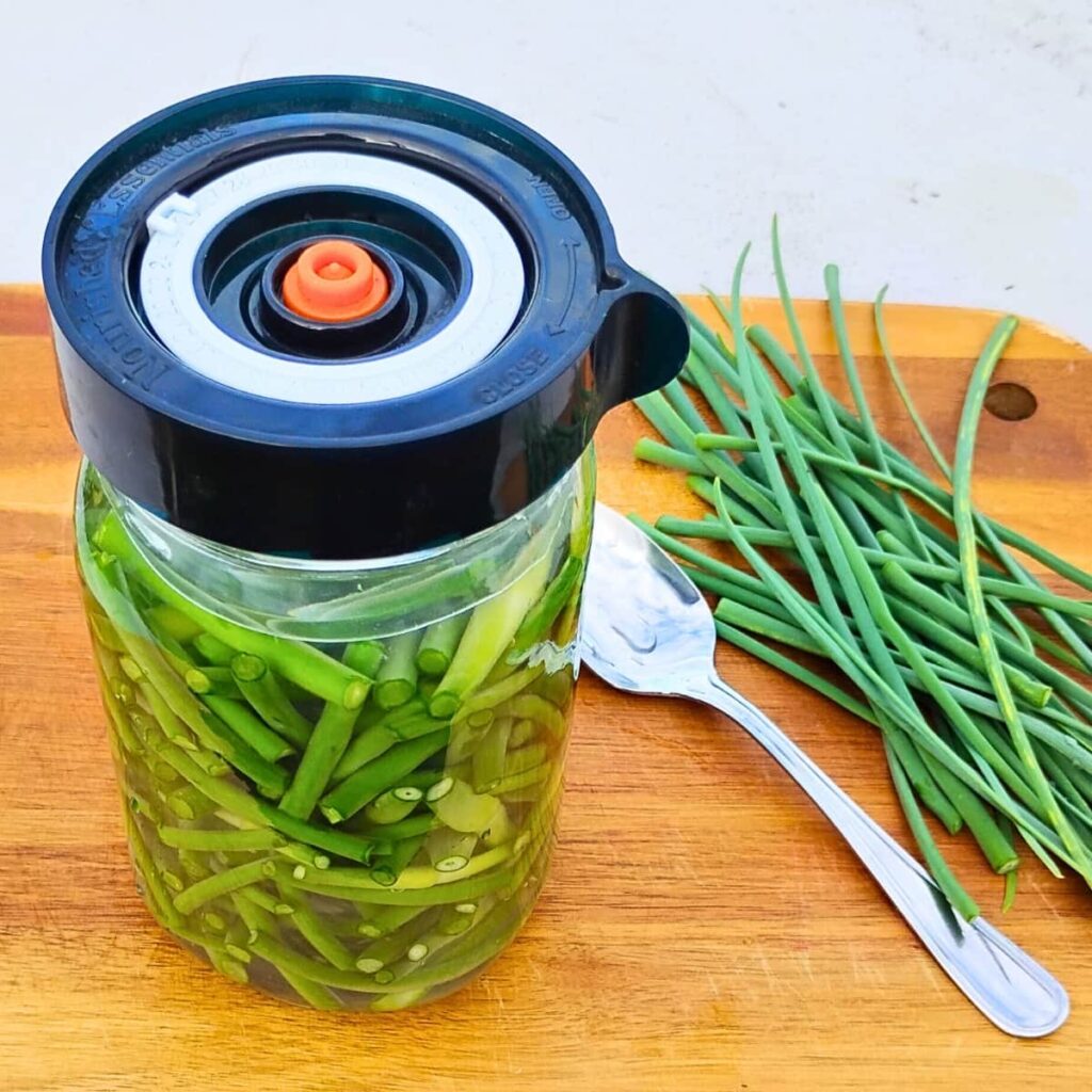 a jar with a fermentation lid that contains garlic scape pieces and brine. there is a spoon next to it and a small bunch of garlic scape tips.