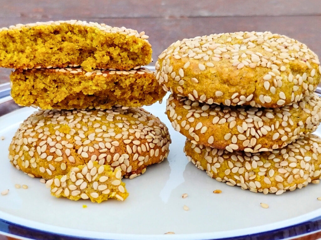 two stacks of lemon sesame tahini cookies on a plate. One of the cookies is broken to reveal the bright interior, colored by honey, ginger bread spices and turmeric.
