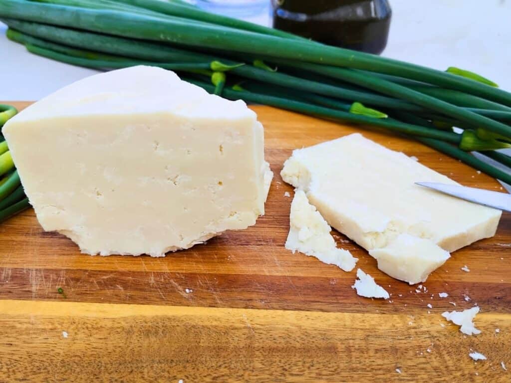 cutting board with a chunk of homemade goats milk cheddar cheese and a slice of it next to it. there are onion flower stalks on the table behind it.