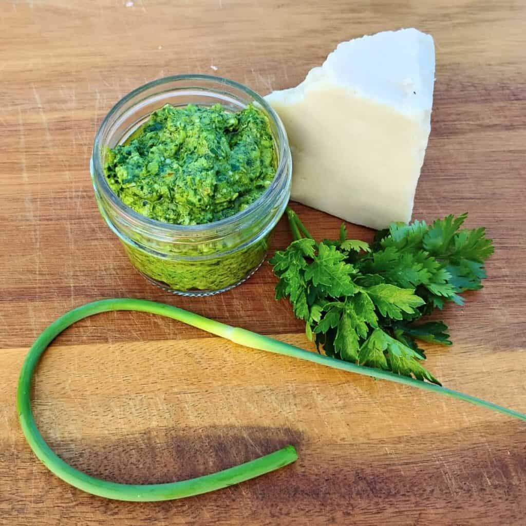 Garlic scape pesto in a small jar on a cutting board with a chunk of cheese , herbs and a garlic scape.