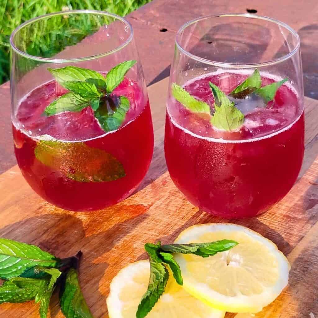 two glasses with ice cubes and fermented hibiscus soda and mint sprigs on a cutting board.
