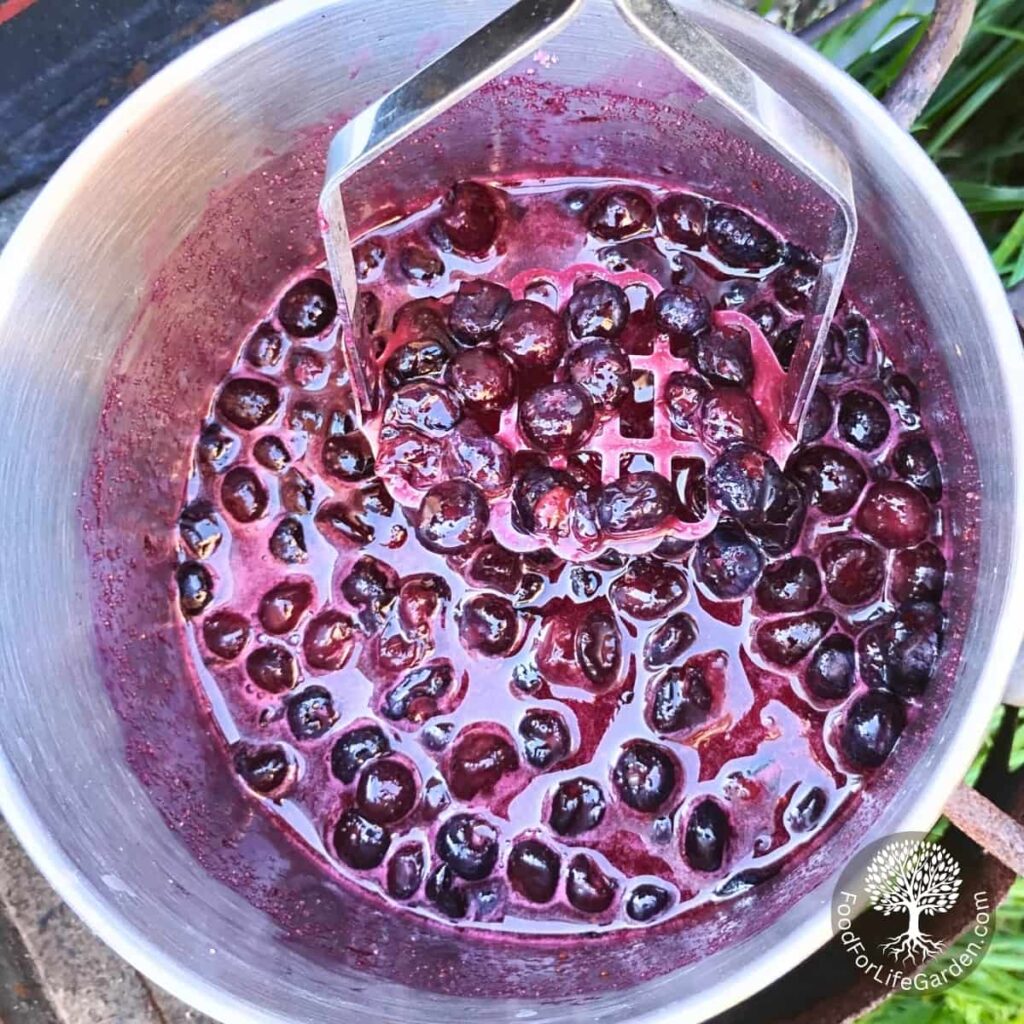 mashing hot blueberries with a potato masher in a pot.