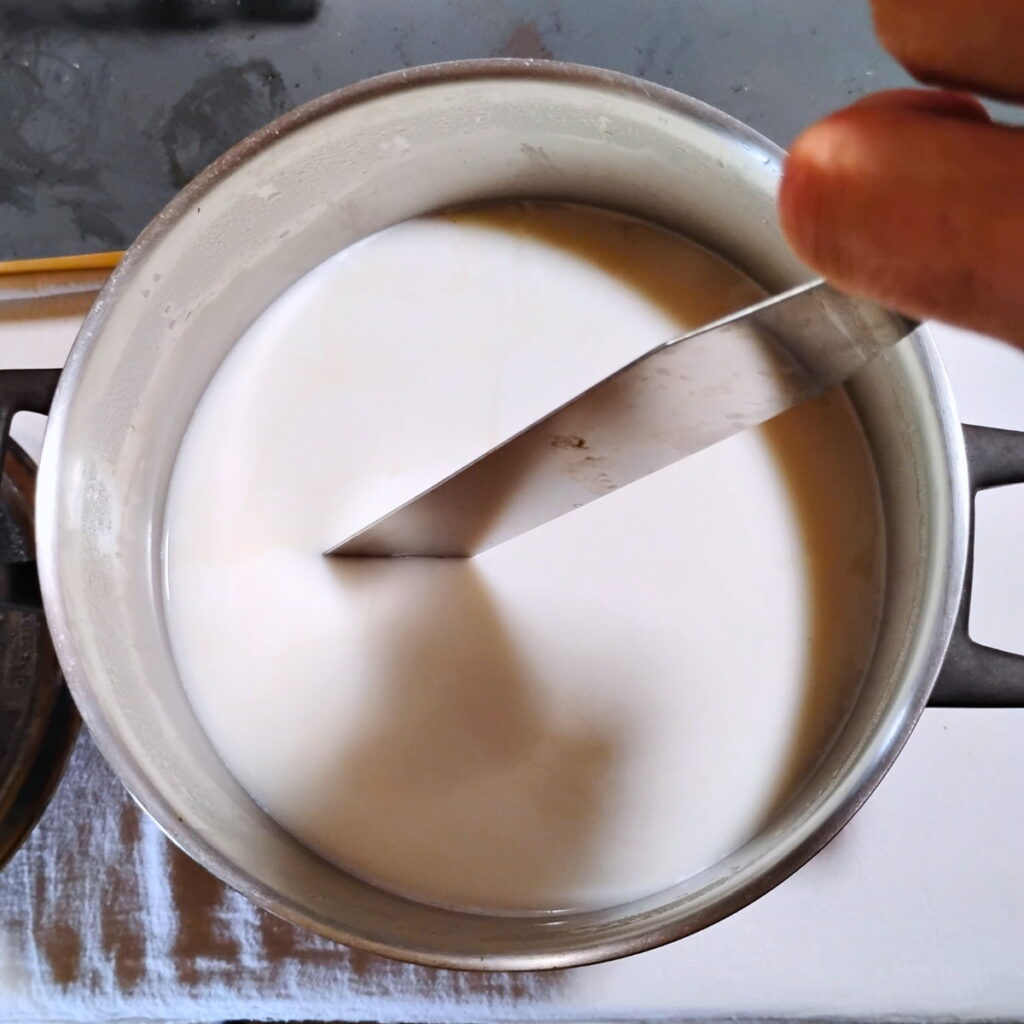 cheese curd is getting cut with a long knife in a pot.