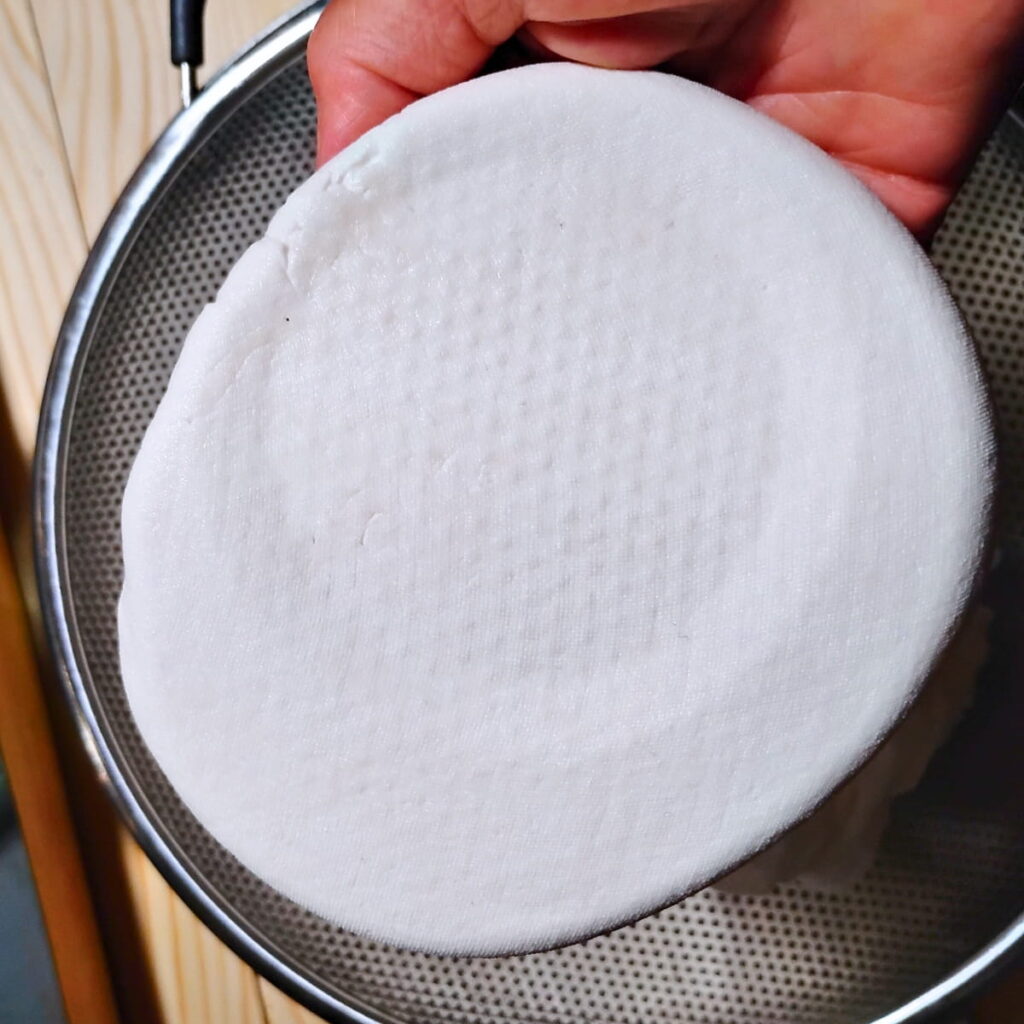 a hand is holding a piece of pressed sirene cheese over a colander.
