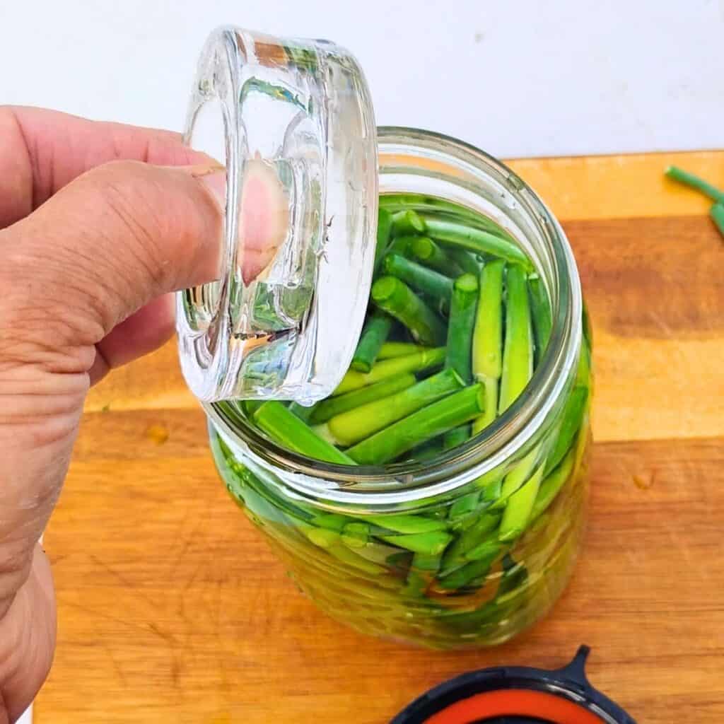 jar full of garlic scapes and brine. a fermentation weight is being placed on top.