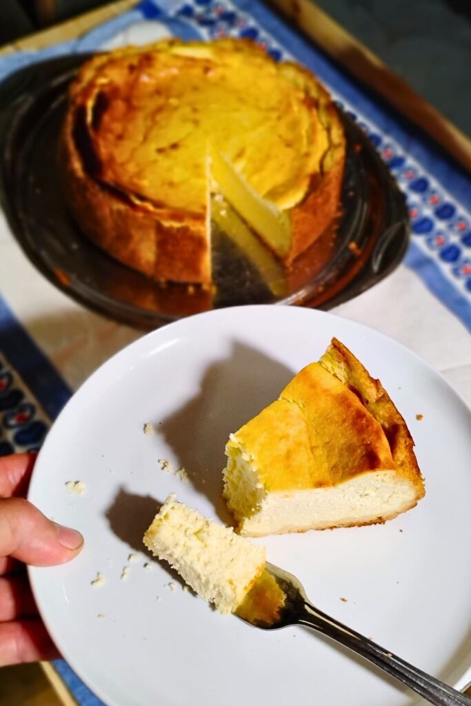 forground shows a slice of German cheese cake or käsekuchen. there is a fork that has a piece on it. Behind it is a full cheesecake with a slice removed.