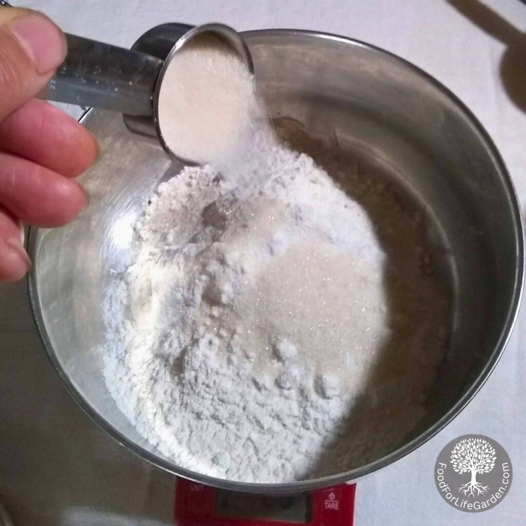 a bowl with flour and a hand is pouring sugar into it from a measuring cup.