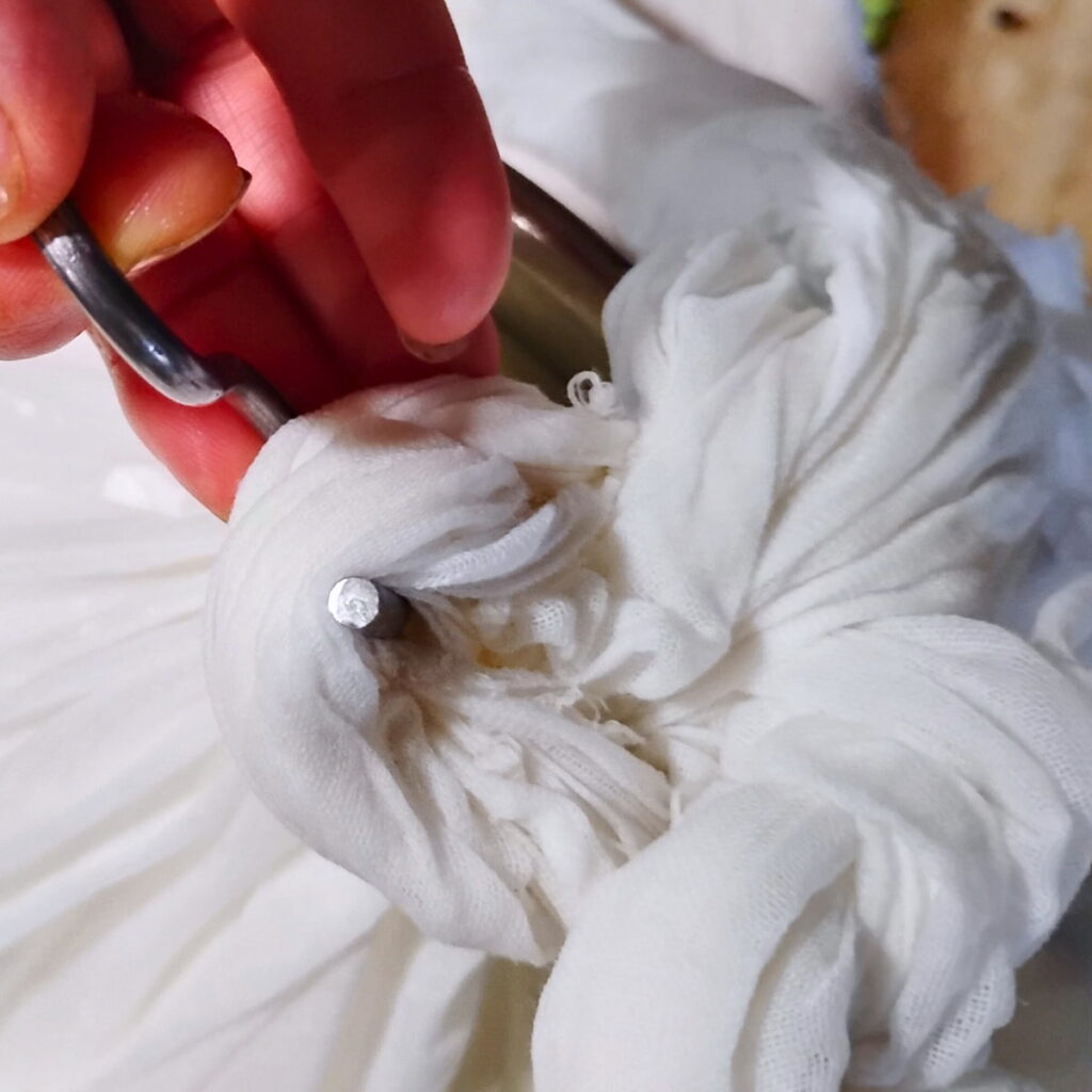 a hand putting a hook through a knot made in the cheese cloth bag.