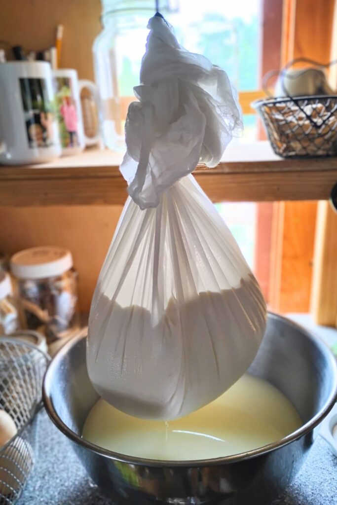 Cheesecurds in a bag of cheese cloth hanging from a hook and dripping whey into a bowl underneath.