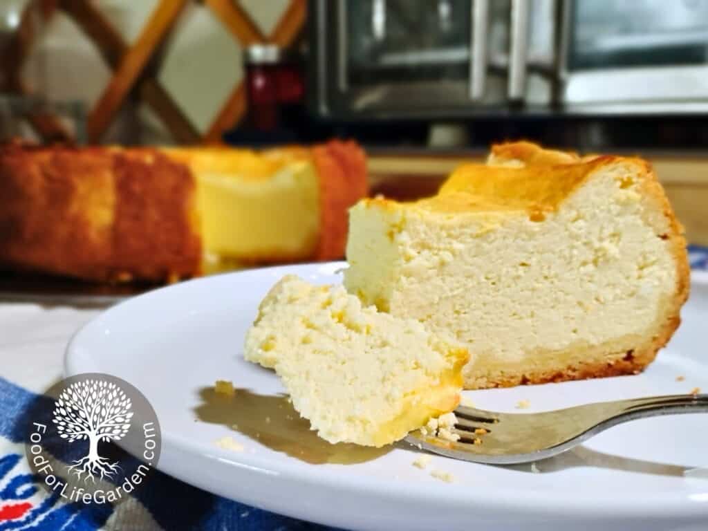 a slice of cheesecake with a fork that has a chunk on it laying in front on a plate. There is a full cake behind it with a slice missing. 