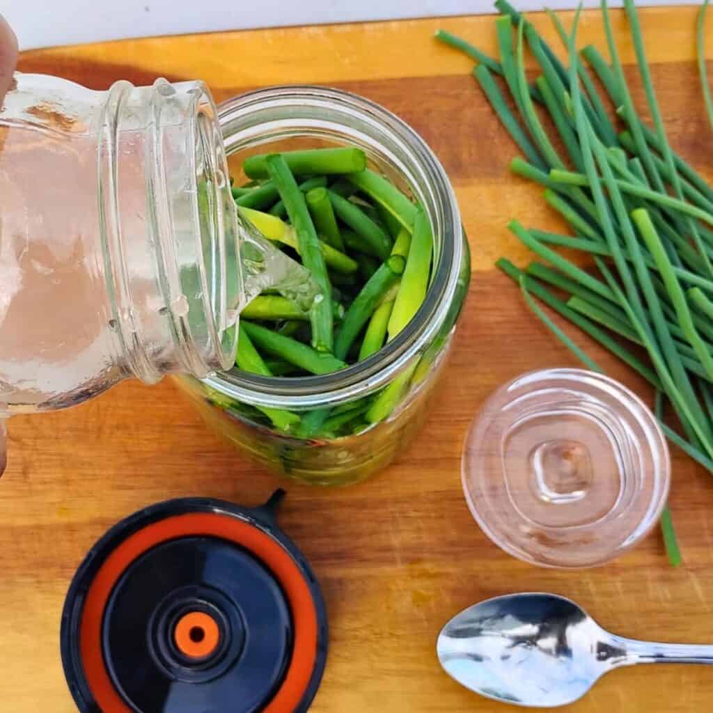 a jar full of garlic scape pieces getting covered with brine from another jar. there is a fermentation weight, a fermentation lid and a spoon next to it, and also some removed garlic scape spikes.