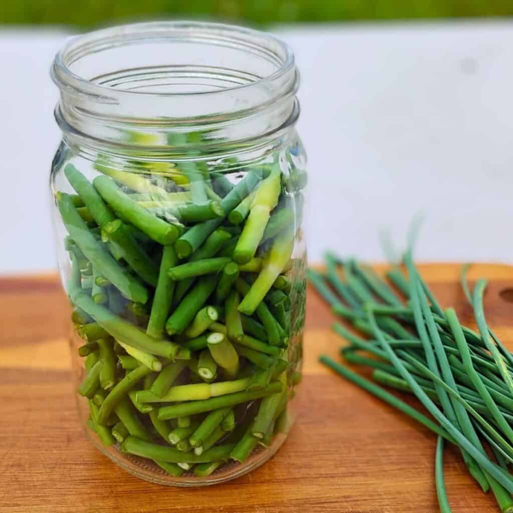 jar full of garlic scape pieces with some flower spikes nest to it on a cutting board.