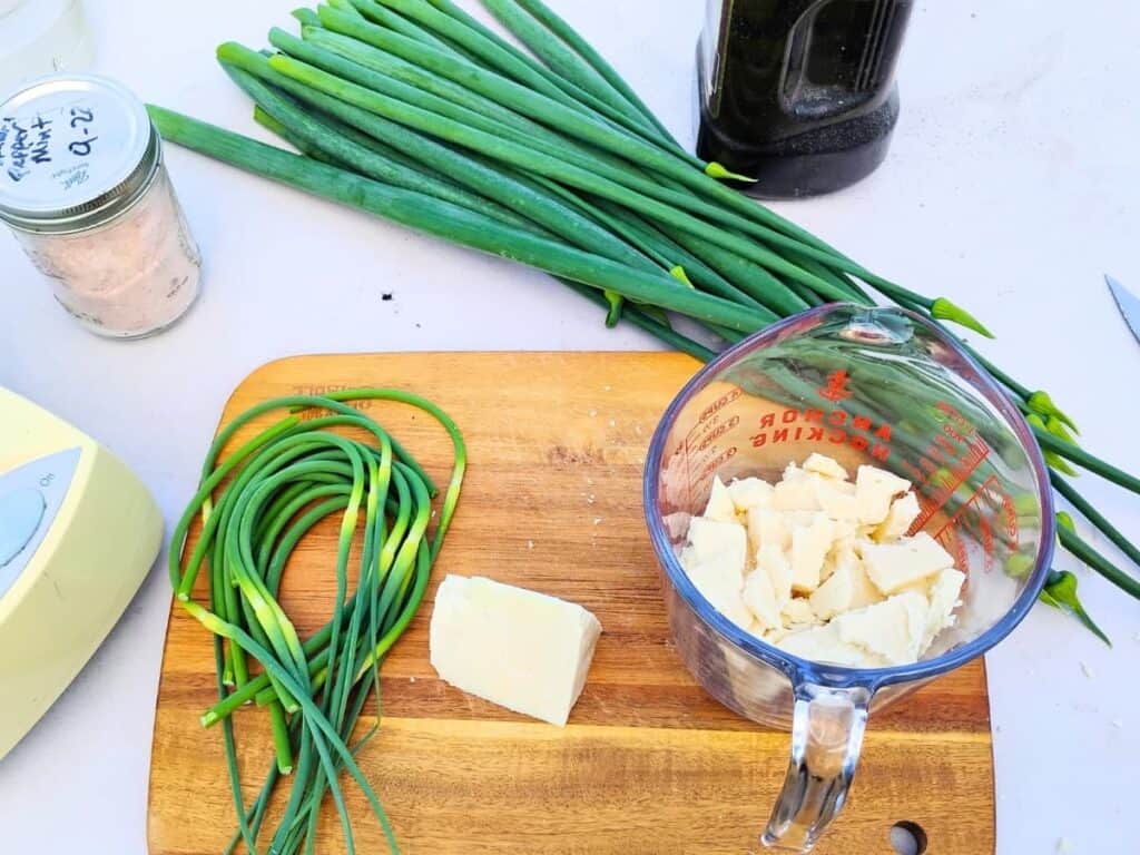 Ingredients for garlic scape and bolted onion greens pesto on a table and cutting board. onion greens, garlic scapes, a jar with salt, a chunk of cheese and some cheese broken up in a measuring pitcher.