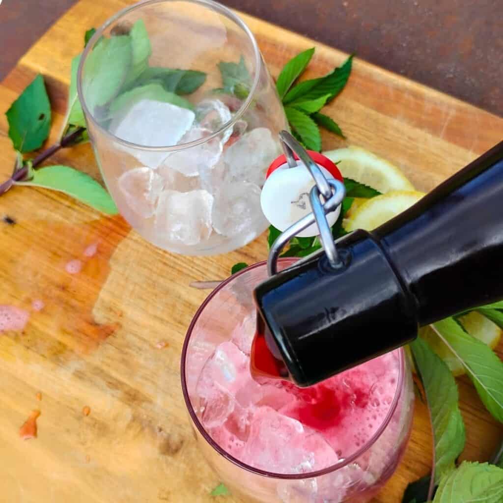 a bottle is pouring fresh brewed hibiscus soda ito a glass with ice cubes. there is another glass with ice cubes waiting to get filled. they are surrounded with mint leaves and lemons.