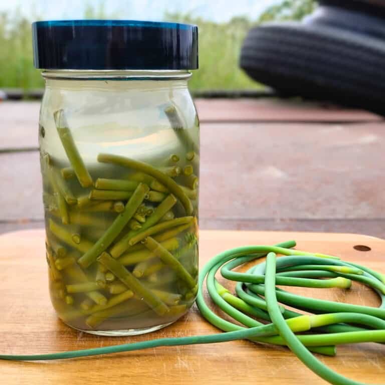 A glass full of garlic scape pieces with a few scapes on the cutting board next to it.