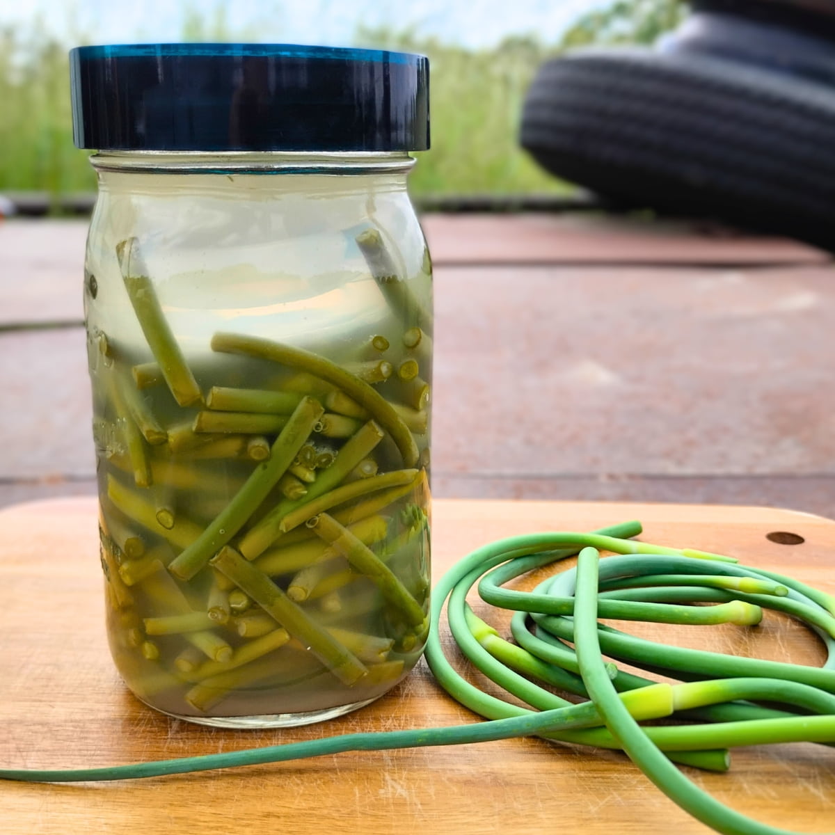 A glass full of garlic scape pieces with a few scapes on the cutting board next to it.