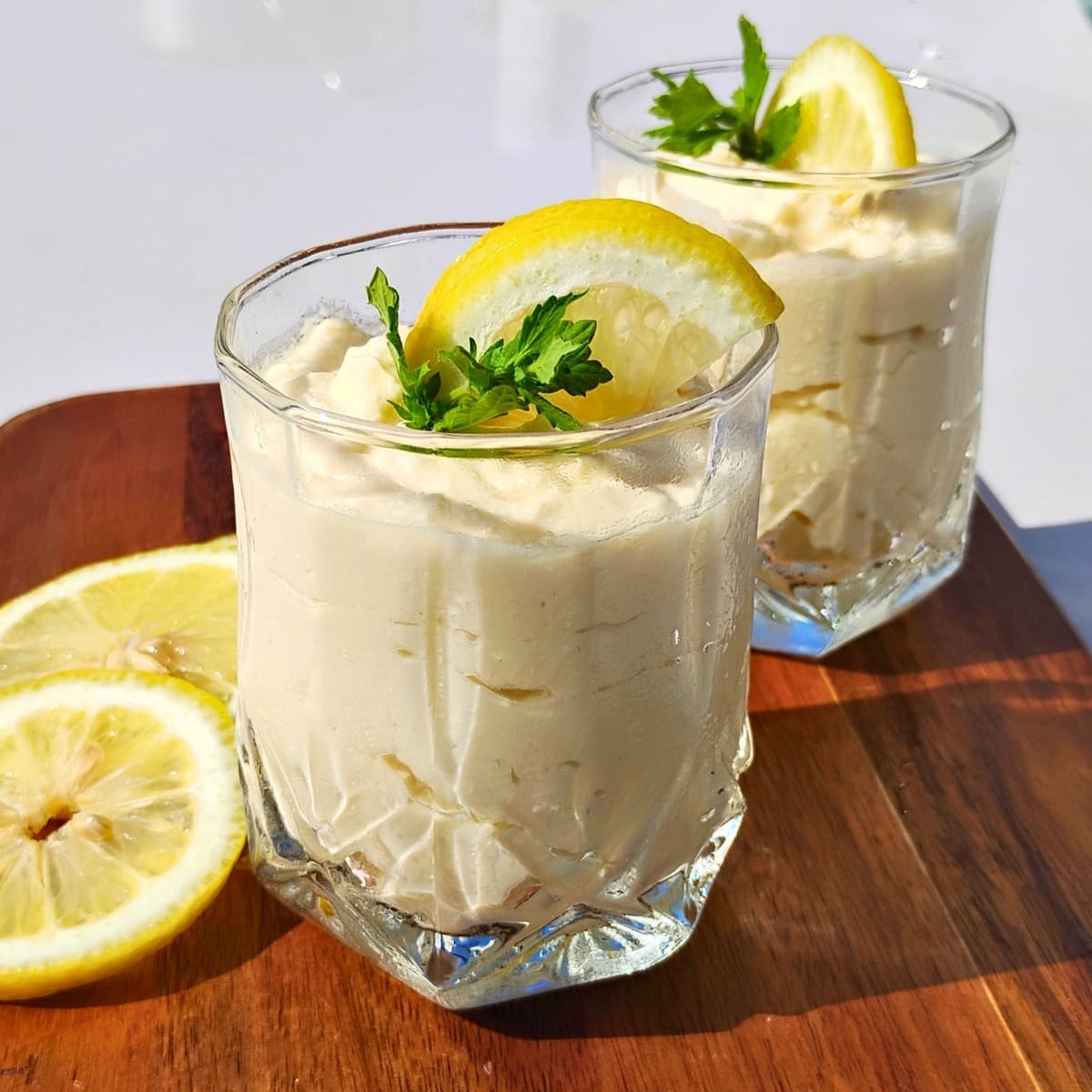 two serving glasses filled with lemon cheesecake mousse and decorated with lemon slices and lemon balm sprigs on a cutting board with more lemon slices.