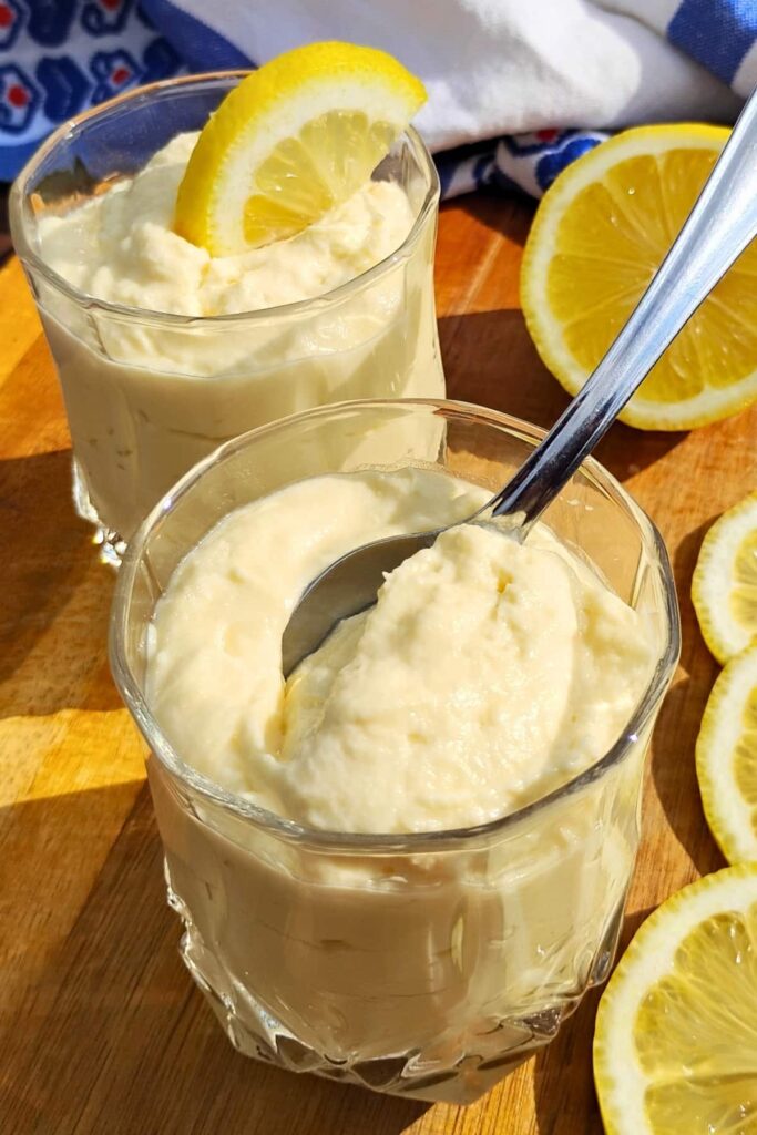 photo shows two serving glasses filled with lemon cheesecake mousse, one decorated with a lemon slice, the other with a spoon in it.