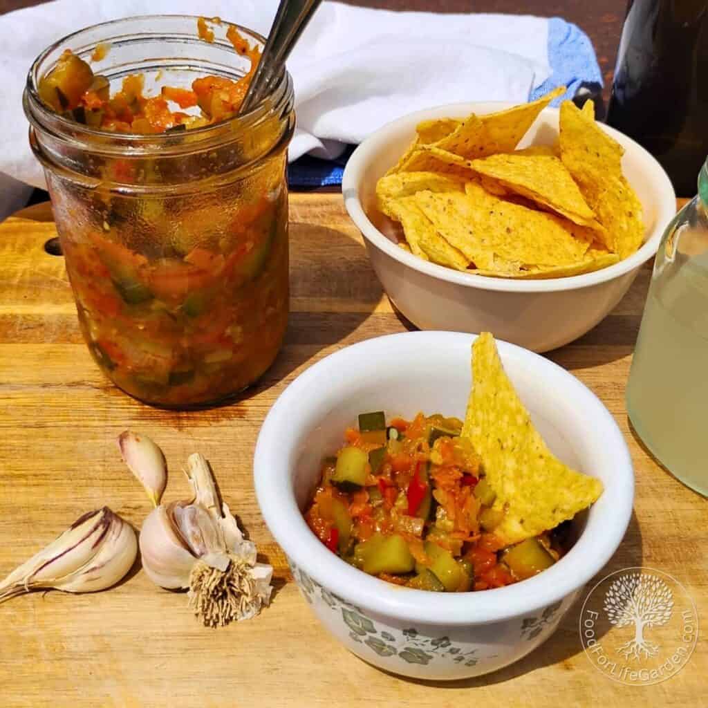 spread of zucchini ikra used as a dip in the front with a corn chip in it. behind it is a bowl filled with more corn chips and beside that a pint canning jar fille with some zucchini vegetable spread.