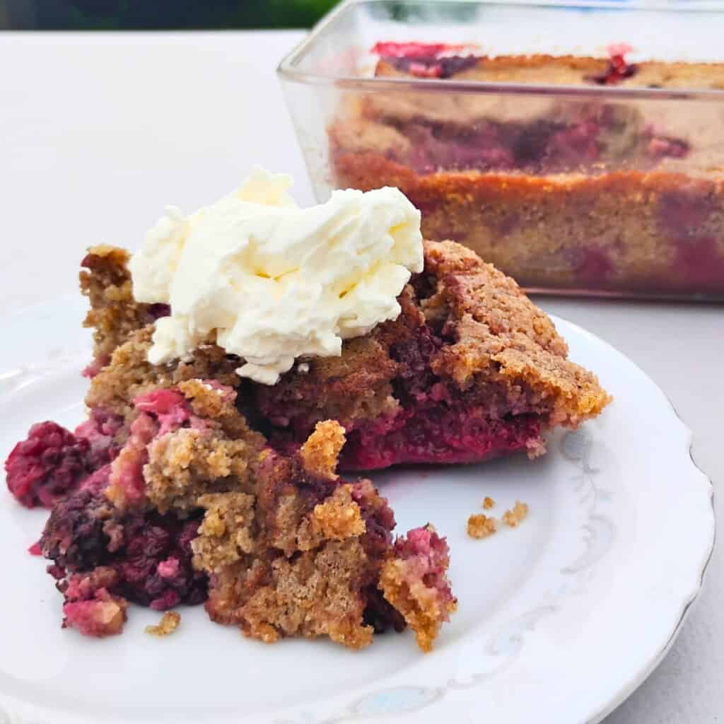 photo of a piece of blackberry cobbler on a plate topped with whipped cream. behind it is baking dish filled with blackberry cobbler.