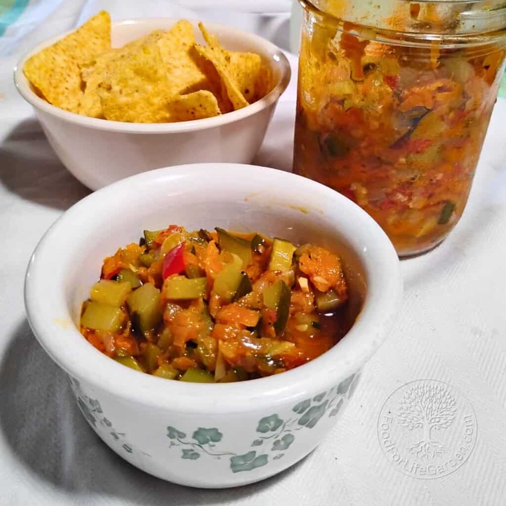 spread of zucchini ikra getting served. a jar with ikra, a bowl in front with some zucchini dip and a bowl with chips behind it.