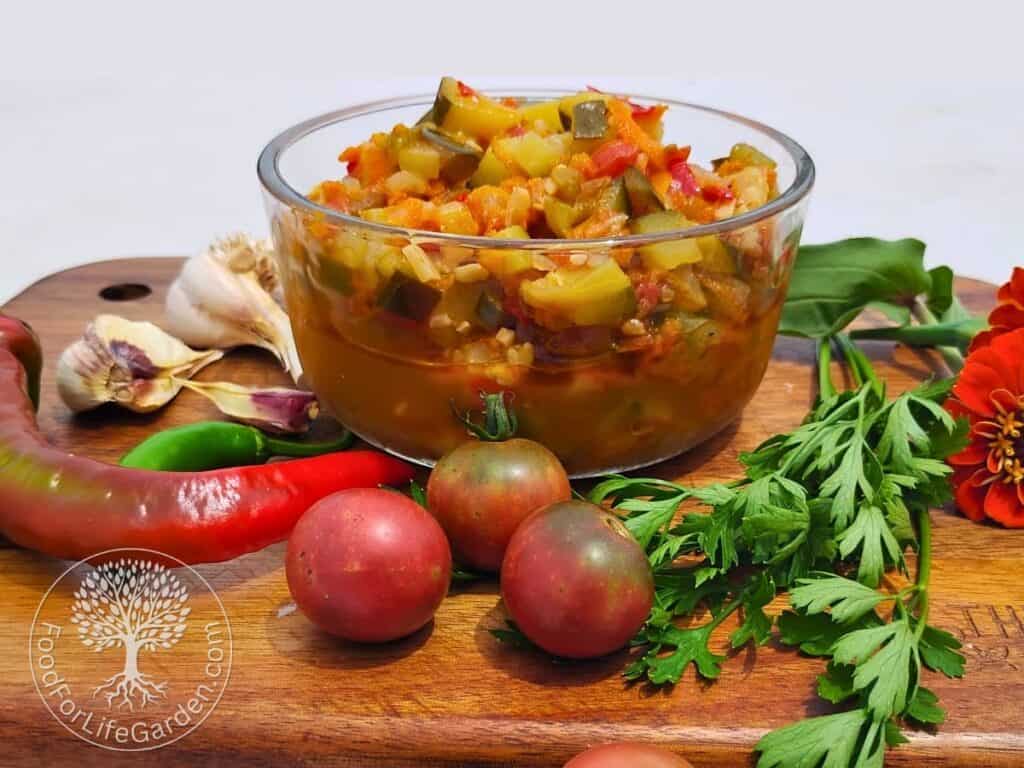 A bowl filled with zucchini ikra, a vegetable spread, dip and sauce, surrounded by some tomatoes, peppers, garlic, parsley, and zinnia and marigold flowers.