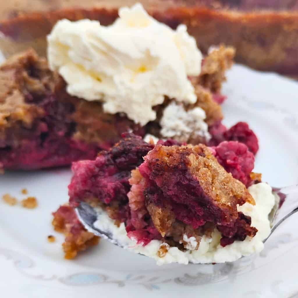 photo of a spoon filled with blackberry cobbler and whipped cream. Behind it is a piece of it on a plate.