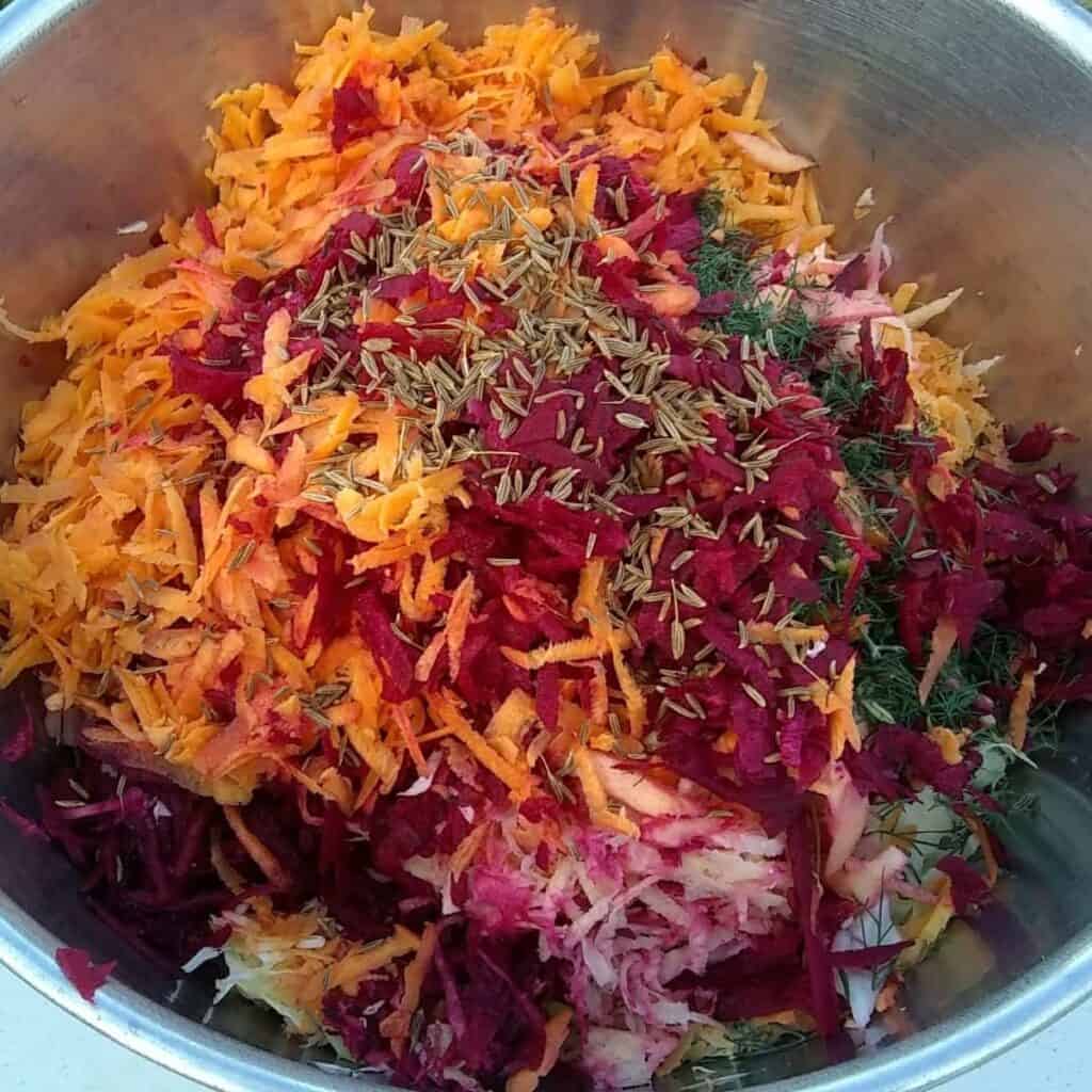 a bowl with prepared ingredients for fermented coleslaw with root vegetables and caraway seeds are sprinkled over the top.
