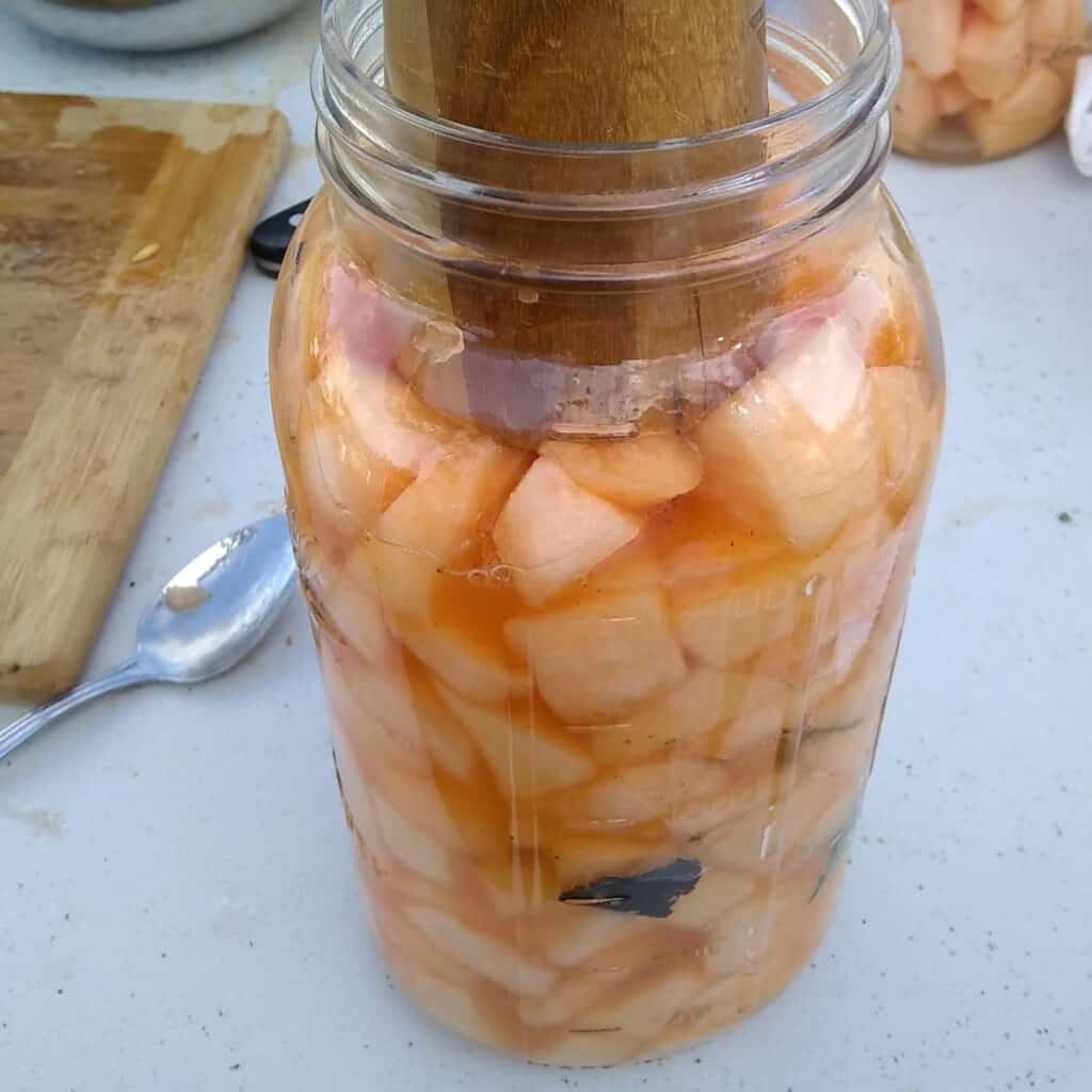 a jar full of ingredients for making fermented cantaloupe and a pickle packer packing it down. 
