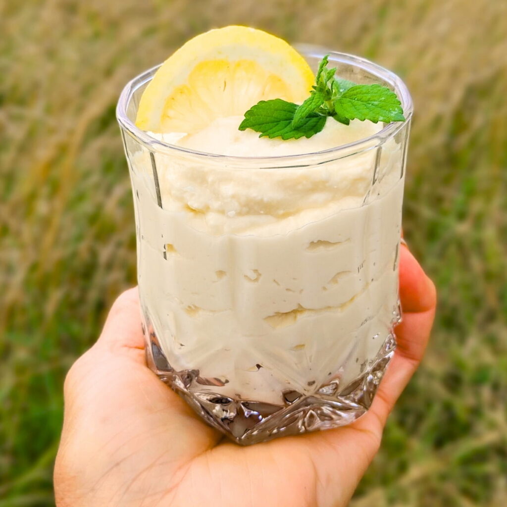 a hand holding up a glass filled with lemon cheesecake mousse, decorated with lemon slice and lemon balm.
