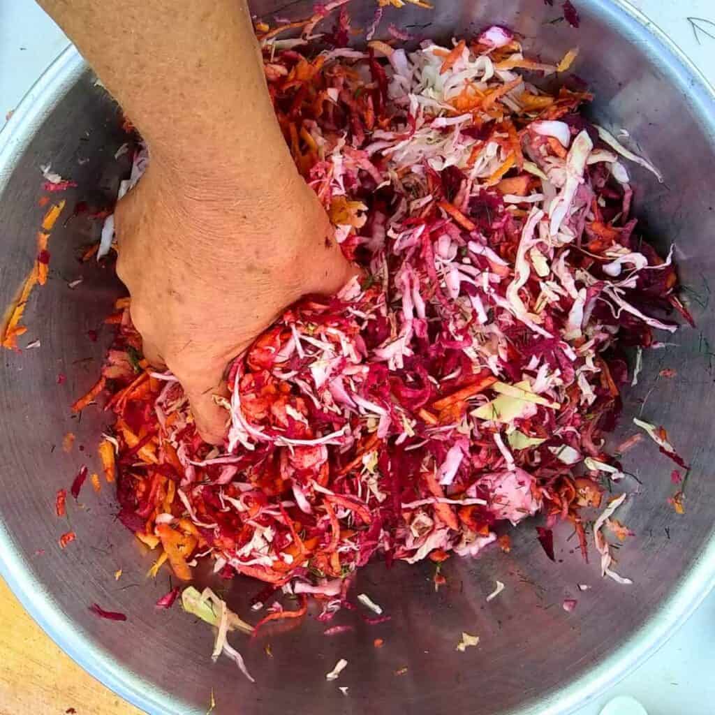 a bowl full of ingredients for fermented coleslaw is kneaded by a hand to create a vegetable juice brine.