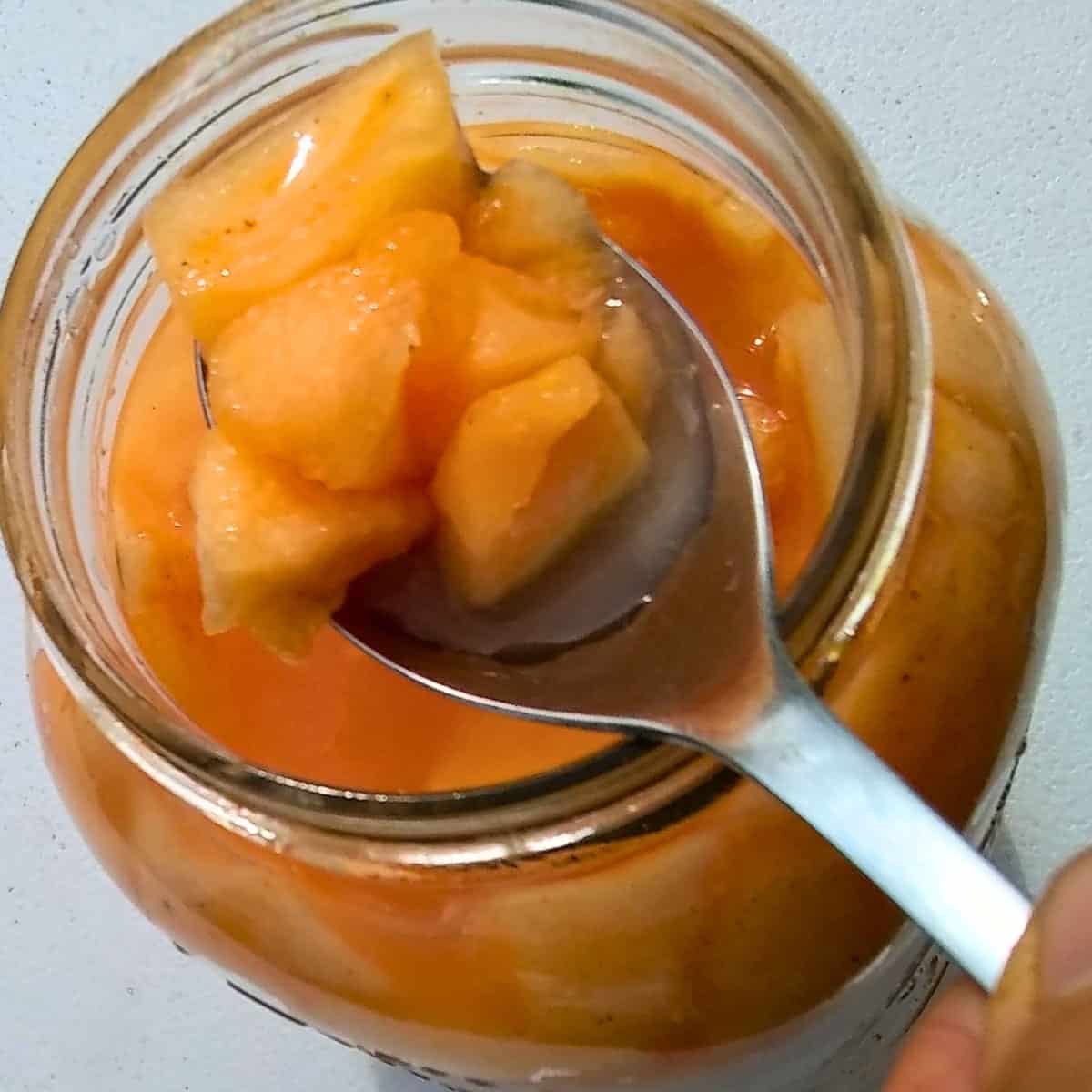 fermented cantaloupe pieces on a spoon, taken from a jar below.