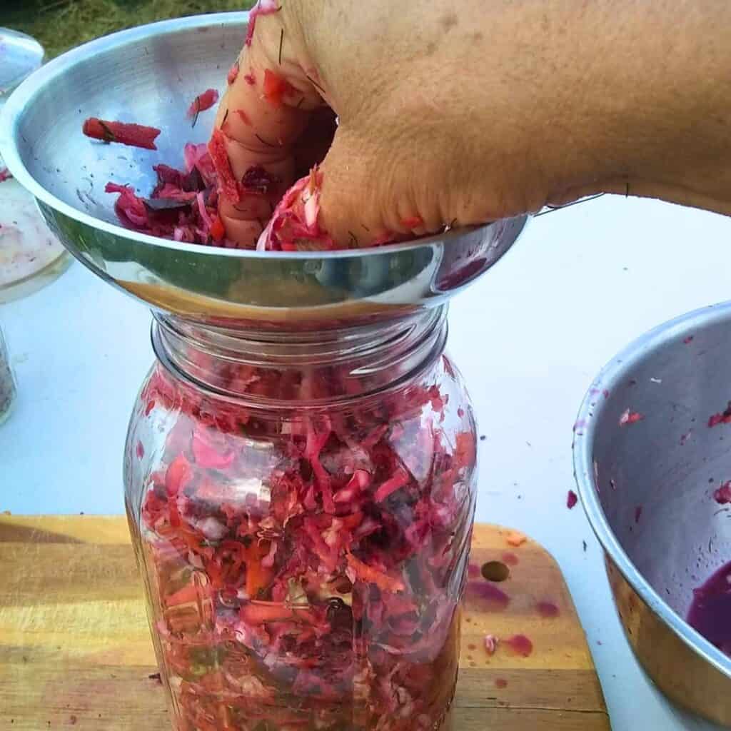 A hand is stuffing a mason jar with ingredients for fermented garden slaw, or coleslaw ingredients.