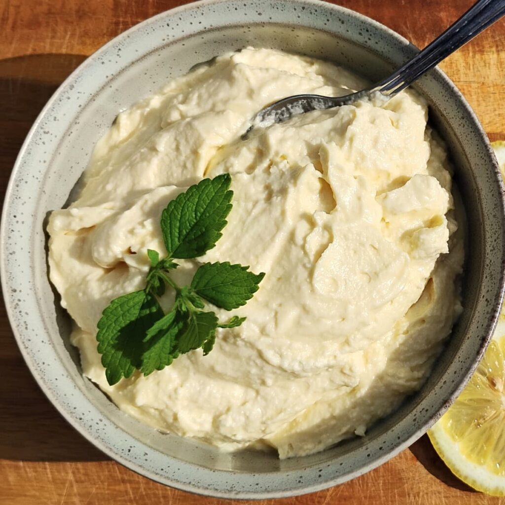 a bowl fllled with lemon cheese cake mousse and topped with a sprig of lemon balm and a spoon in it.