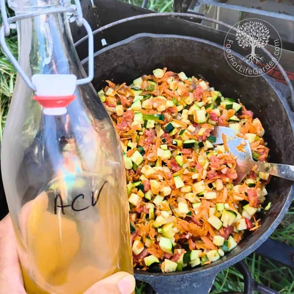 cast iron dutch oven with ingredients getting added and a hand holding a bottle of vinegar that has the letters ACV on it.