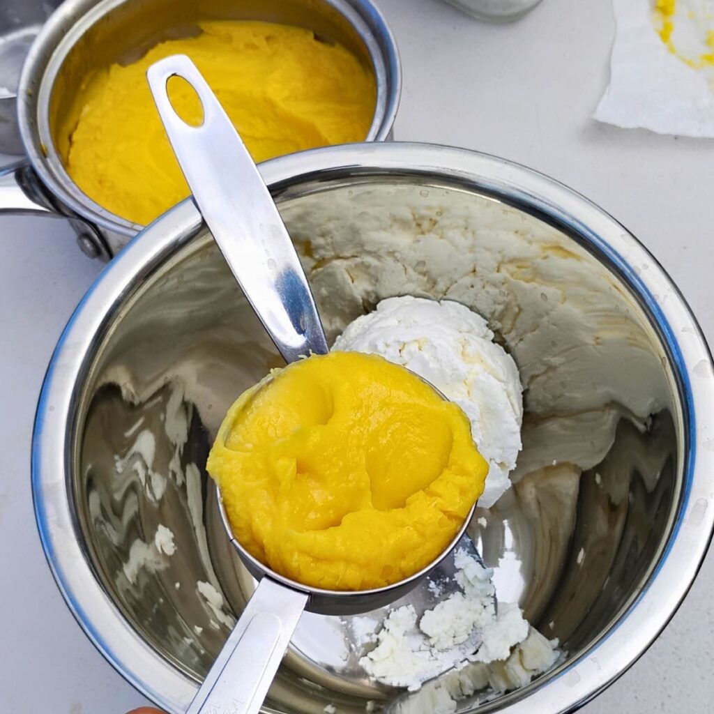 photo of a bowl with some creamy cheese and a measuring cup filled with lemon curd that is getting added to the cheese. there is a pot filled with lemon curd behind it.