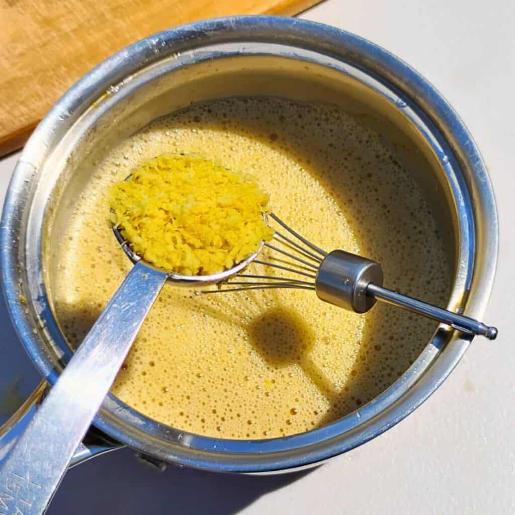 lemon zest is getting added with a spoon to a pot that has blended lemon curd ingredients.