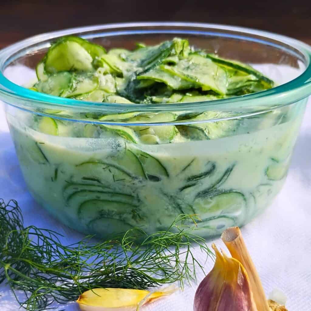 photo shows the sideview of a bowl filled with creamy cucumber salad and a dill sprig and some garlic cloves in the foreground.