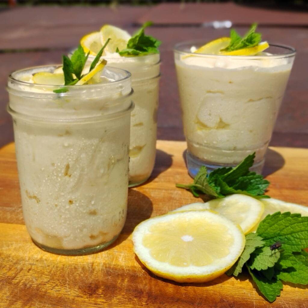 photo shows three glasses filled with lemon cheesecake mousse on a wooden board all decorated with lemon slices and lemon balm.