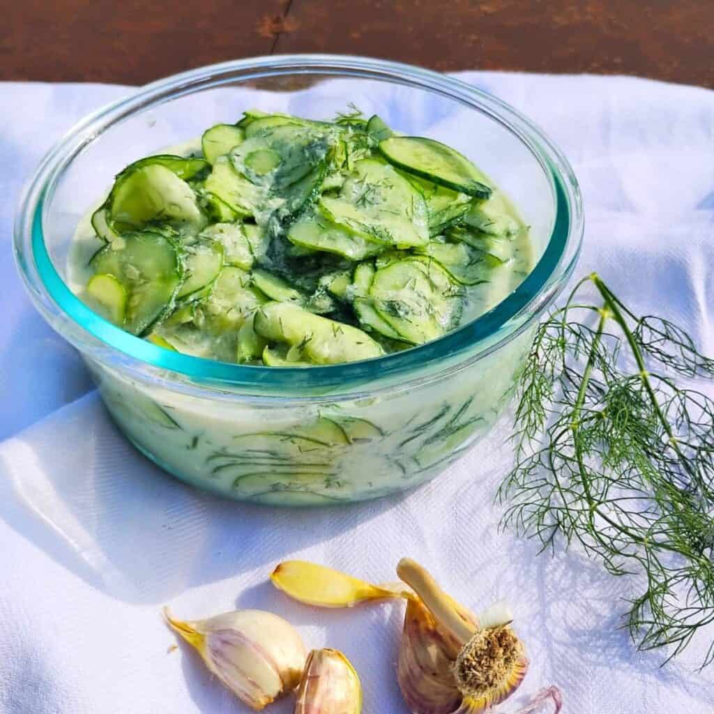 A bowl filled with creamy cucumber salad and some dill and garilc around it.