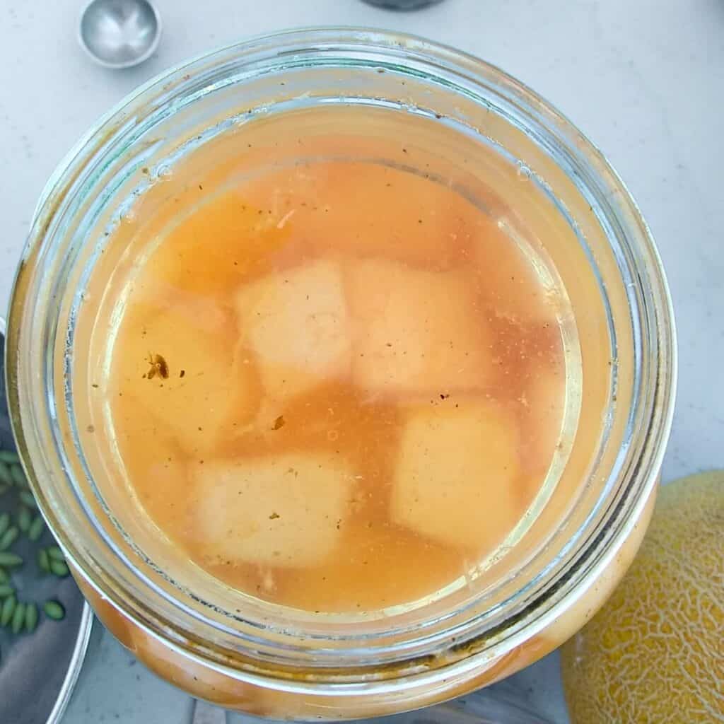 a jar filled with cantaloupe pieces and brine with a glass fermentation weight on top. viewed from above.